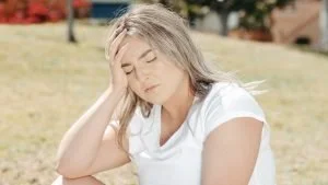 A woman experiencing headaches sits on the grass with her hands on her head.