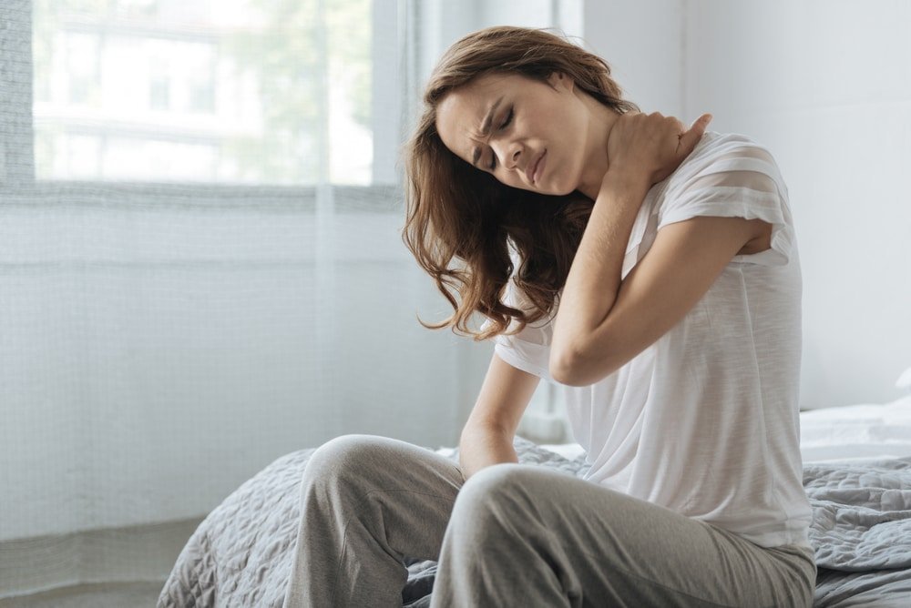 A woman in London is sitting on a bed with a pain in her neck.