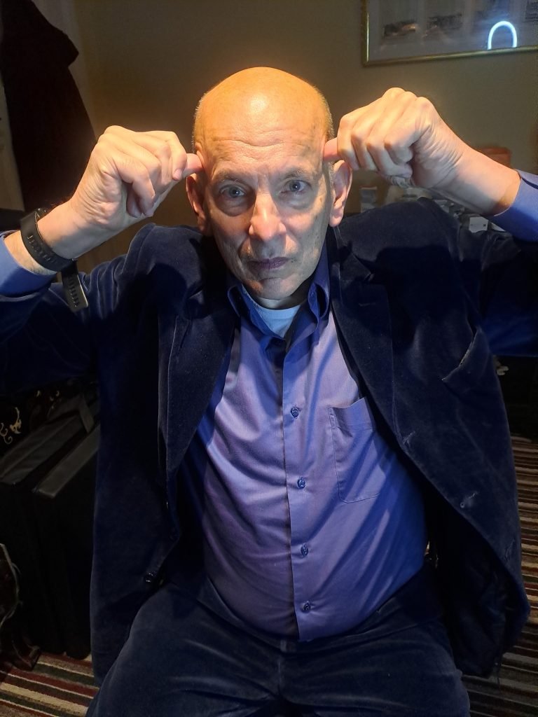 A bald man in a blue jacket with his hands on his ears.