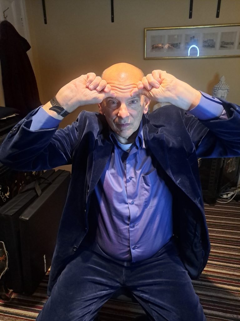 A bald man sitting in a room with his hands up.