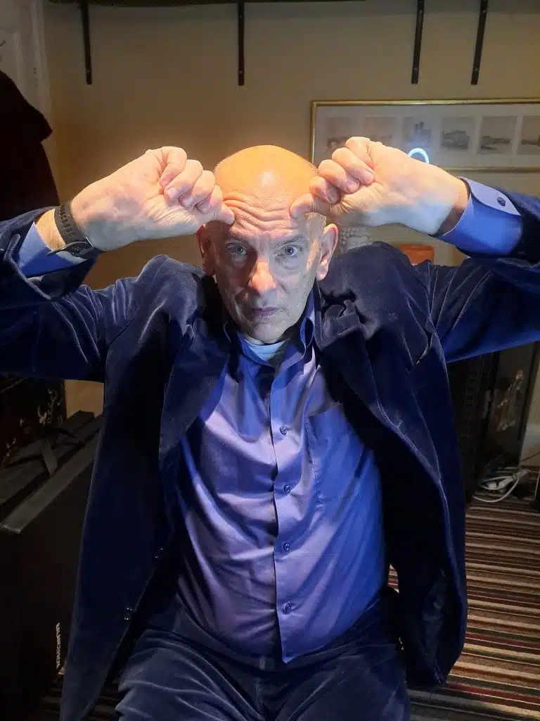 A bald man in a blue suit with his hands up.