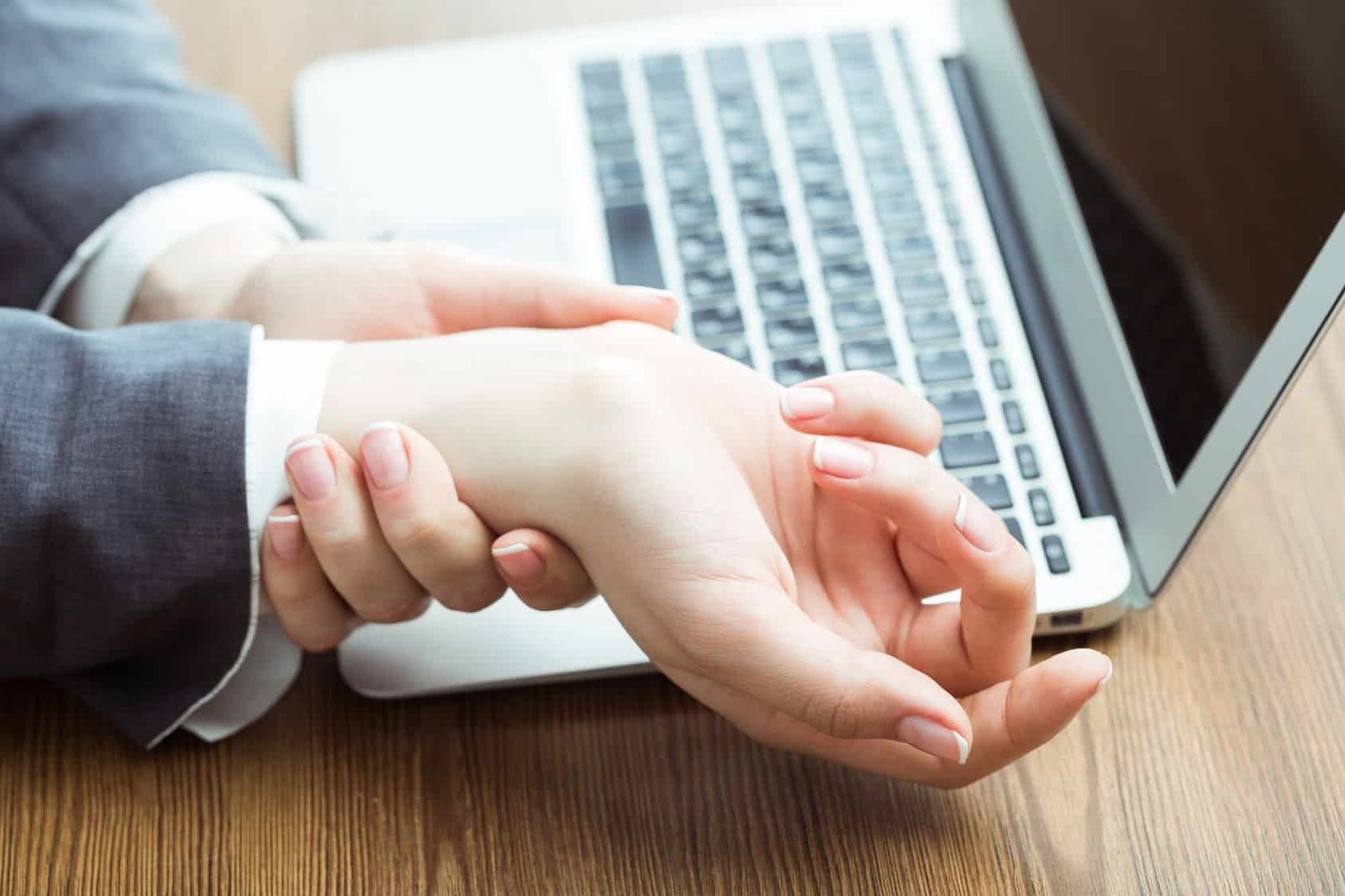 Person in a gray suit holding their wrist in front of an open laptop on a wooden desk.