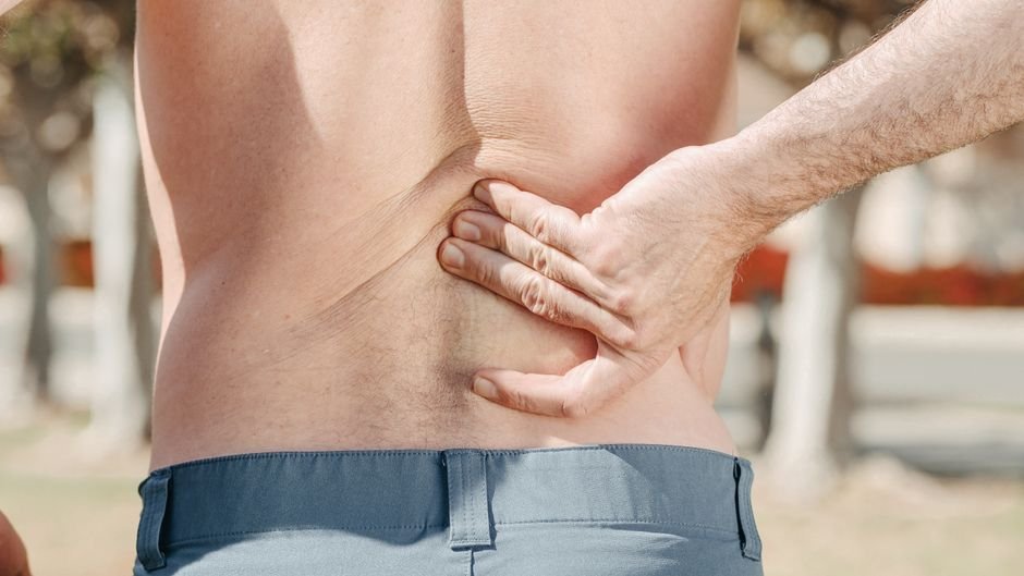 A person outdoors holds their lower back with one hand, appearing to experience discomfort or pain.